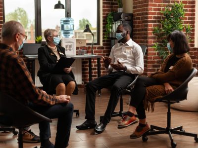 Multi ethnic people discussing about addiction at support group therapy, during covid 19 pandemic. Patients with face masks at aa meeting to cure mental health problems with specialist.