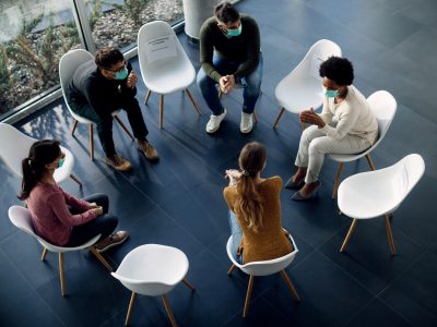 High angle view of group therapy participants sitting in a circle and talking while wearing protective face masks due to COVID-19 pandemic.